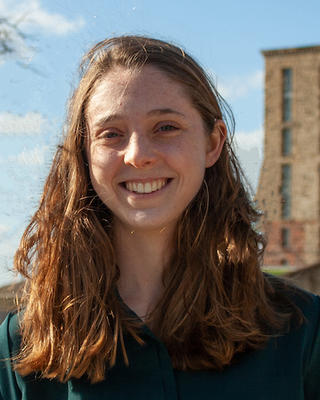 Catriona Breen with long hair and a green shirt, in front of blue sky, brick building, and grassy area