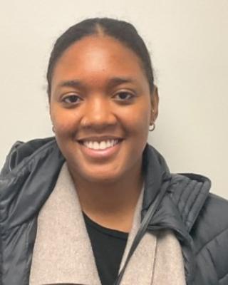 Chaolyn Clay, smiling, dark hair pulled back, wearing dark grey coat, light grey scarf, in front of beige background