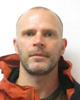 Chris Preheim with very short hair and beard, orange and black coat, white background