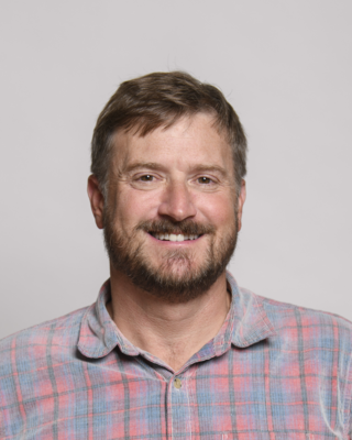 Christian Flynn smiling, short brown hair and beard, red and blue plaid shirt, grey background