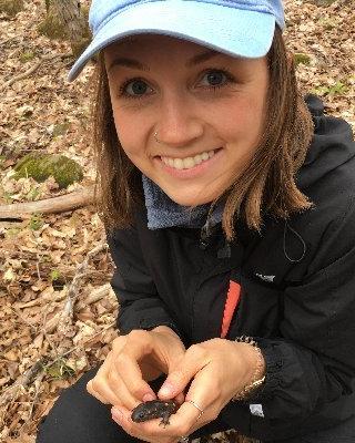 Christina Steinecke with long brown hair, squatting on leaf-littered ground, wearing dark outdoor clothing and a light blue hat, holding a small dark amphibian