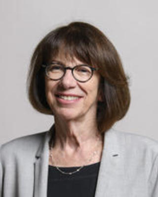 Colleen Cavanauth with medium dark hair wearing glasses, a black top and grey jacket in front of grey background
