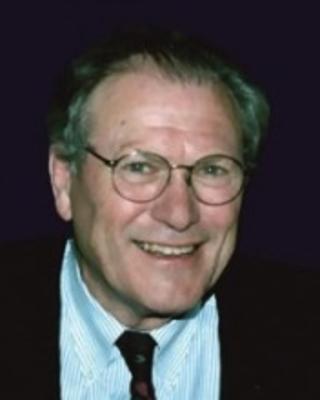 Alfred Crompton smiling, wearing glasses, a dark jacket, white button down, and dark tie, in front of dark background