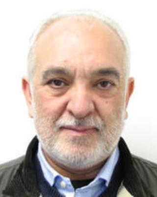 Daniel Sanchez  whit short white hair and beard, blue shirt, dark coat, in front of white background