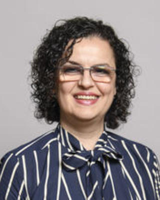 Diana Gjino with medium lenght brown curly hair, wearing glasses and a blue shirt with white stripes and bow collar