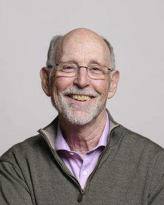 Don Pfister smiling, with short grey hair and beard, glasses,  pink button down, and taupe quarter-zip