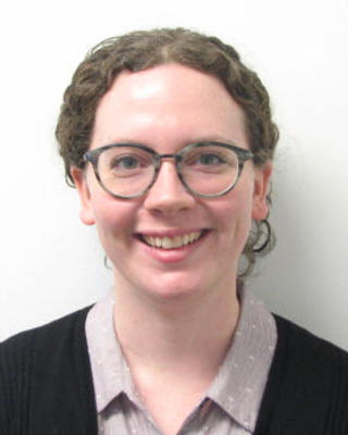 Emily Reynolds smiling, with glasses and curly medium hair pulled back, wearing light button-down and black cardigan