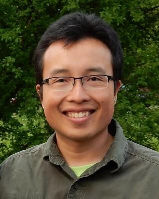 Evan Hoki with short dark hair, glasses, and a muted green shirt, in front of green leafy trees