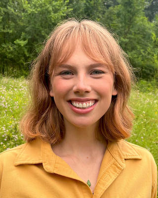 Eve Farrell with medium length red hair and bangs, wearing a yellow shirt, in front of trees and a flowering meadow