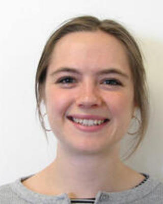 Grace Burgin, dark blonde hair pulled back, wearing a grey cardigan, in front of a white background