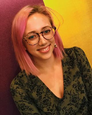 Grace Pisano with long pink hair, glasses, and green and black floral shirt, in front of magenta and yellow wall