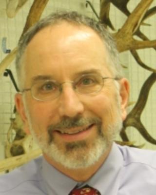 James Hanken, with short grey hair and beard, wearing glasses and smiling in front of a wall of antlers