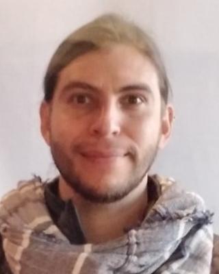 Javier Luque with light brown hair and a striped scarf, in front of a light grey background
