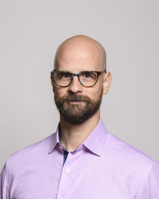 Javier Ortega-Hernandez wearing colorful glasses, a pink check shirt, and with a short dark beard, in front of grey background