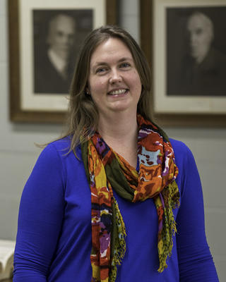 Jeannette Everritt with long dark blond hair, bright blue shirt and colorful scarf, wall with framed black and white portraits behind her