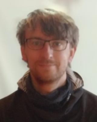 Jonas Geburzi with short brown hair, wearing glasses and a dark coat, in front of a beige background