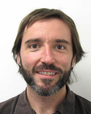 Juan Losada with brown hair and dark beard, wearing a brown shirt, in front of a white background