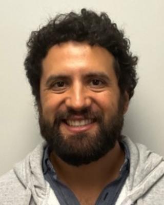Juan Ramirez Lerma smiling, with cruly dark hair and beard, mustache, wearing medium grey shirt and light grey hooded sweatshirt
