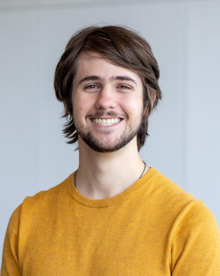 Julius Tabin with brown hair and beard, wearing a dark yellow sweater, in front of a light grey background