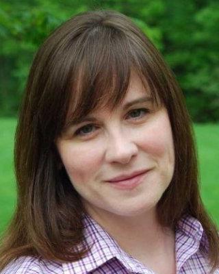 Kathleen Pritchett-Corning, with long brown hair and bangs, wearing plaid purple and white shirt, green grass and shrubs in background