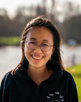 Kaylin Chong with long dark hair and glasses, wearing a dark top, in front of water with grassy embankments with trees