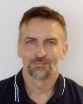 Ken Pero with short brown hair, dark polo with white stripes on collar, grey background