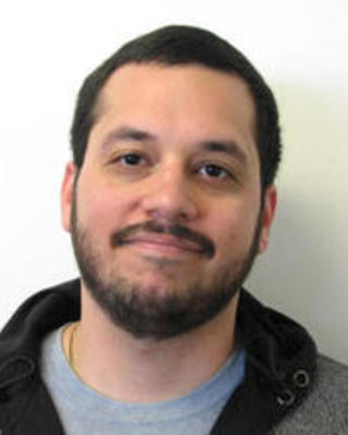 Kevin Laboy Juarez with short dark hair and beard, waring a grey shirt and fleece, in front of a white background
