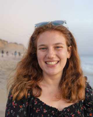 Kristen Saban with long red hair, black top, sunglasses on head, in front of sandy beach and grey sky