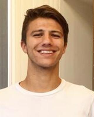 Landen Gozashti with short brown hair and a white top, standing in front of beige interior wall