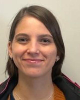 Leticia Rezende de Oliveira with medium length brown hair, an orange sweater and black coat, in front of beige background