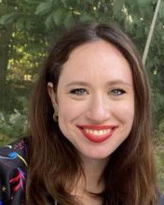 Lily Shapiro with long brown hair, black shirt with bright pattern, in front of leafy trees