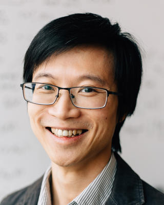 Louis Fan with short black hair, wearing glasses, a striped shirt, and dark blazer, in front of mottled light grey background