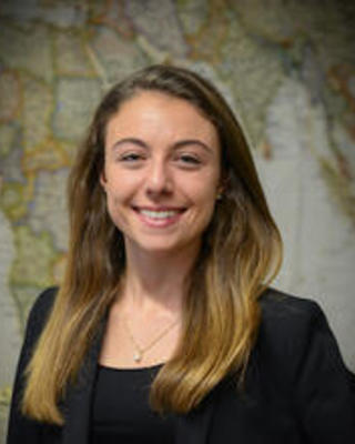 Lucrecia Aguilar with long light brown hair, wearing dark top and blazer, in front of world map