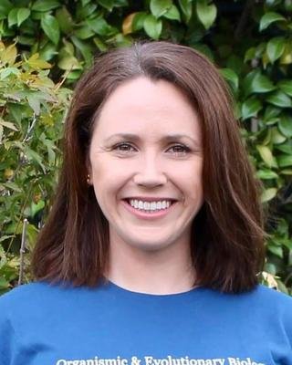 Lucy Wilson with shoulder length brown hair, wearing blue OEB shirt, in front of green bushes