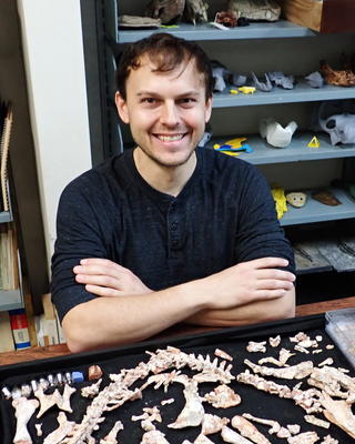 Mark Wright with short brown hair and a dark shirt, sitting with arms crossed, a table full of fossilized bones in front of him, shelves of fossils and models behind him