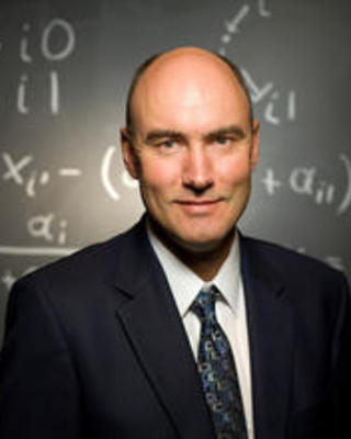 Martin A. Nowak wearing a suit and tie, in front of a chalkboard with equations on it