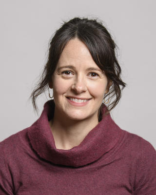 Melissa Aja with brown hair, wearing a red top, in front of grey background