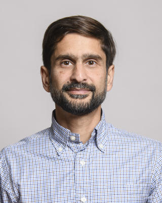 Michael Desai with short dark hair and beard, wearing a blue check button down, in front of grey background