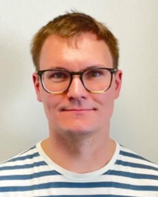 Moritz Weglage with short red hair, glasses, blue and white striped shirt, in front of beige background