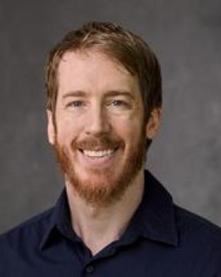 Patrick Gorring with short light red hair and beard, wearing a dark blue top, in front of a grey background