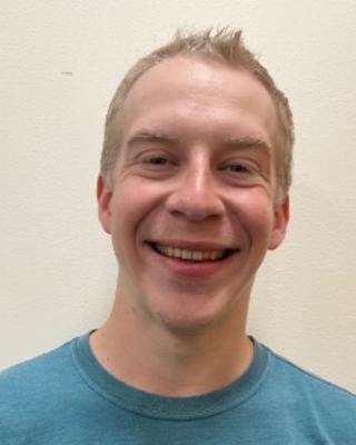 Patrick McKenzie with short blond hair wearing a teal shirt, in front of abeige background