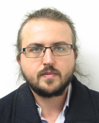Pawel Zmarz with light brown hair and beard, glasses, and dark coat, in front of white background