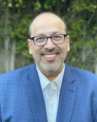 Peter Girguis wearing blue blazer, white shirt and glasses, in front of leaf covered building