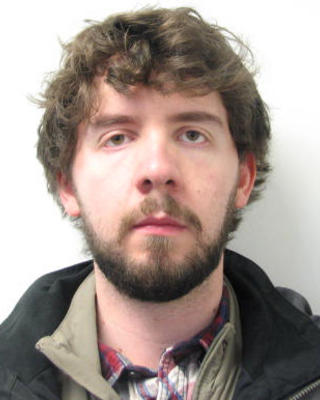 Robert Brocklehurst with short, curly brown hair and beard, wearing layered shirts and dark coat, in front of white background