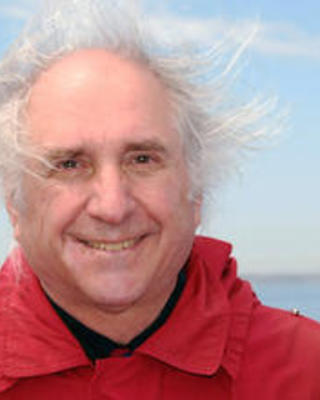 Robert M. Woollacott with windblown grey hair, red jacket, blue sky and water in background