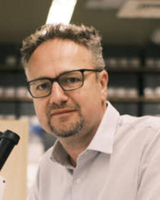 Rudy Lerosey-Aubril with short brown hair and goatee, dark glasses, white button down shirt, next to microscope