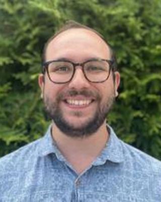 Ryan Schmidt with brown hair and beard, wearing patterned blue shirt, in front of green bushes