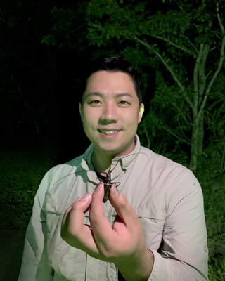 Sang Il Kim wearing a khaki shirt, in front of a tree in the dark, holding a large beetle toward the camera
