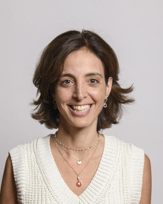 Sarine Der Kaloustian, smiling, with short brown hair, cream sleeveless sweater, grey background