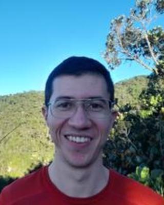 Sebastian Martinez-Salazar with short brown hair, glasses, and a red shirt, in front of green mountainside and blue sky